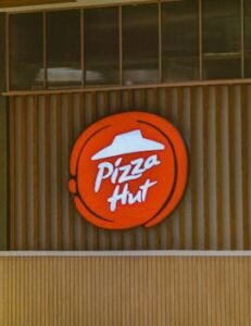 Illuminated Pizza Hut sign on a wooden storefront captured in daylight.